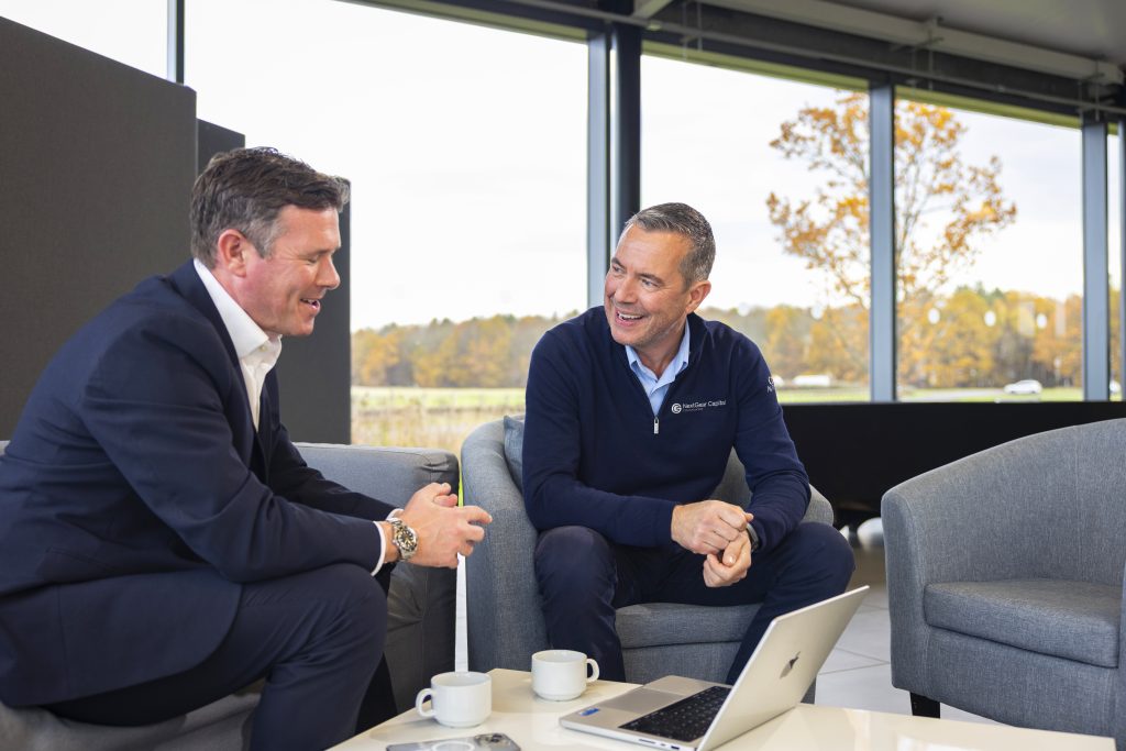 NextGear Capital employee wearing a dark blue jumper is sat with a customer, talking and smiling.