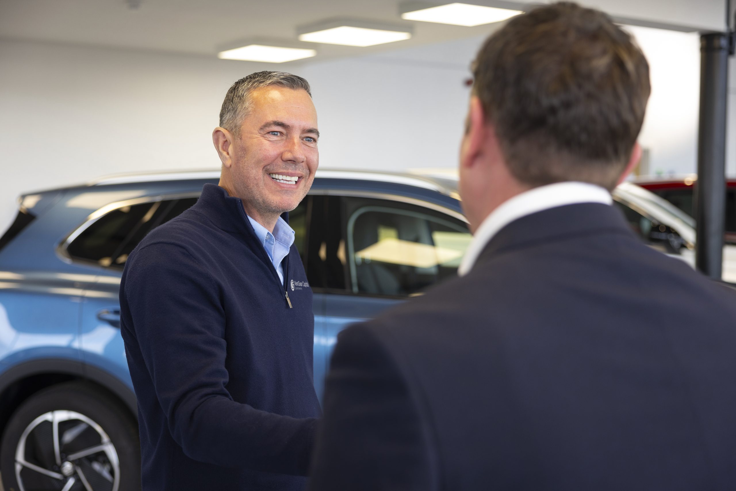 NextGear Capital employee wearing a dark blue jumper shaking hands with a customer with a light blue car in the background.