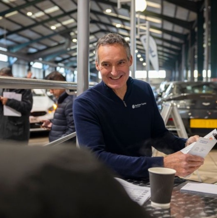 A NextGear Capital employee wearing a blue NextGear Capital jumper smiles at a customer in an auction room.